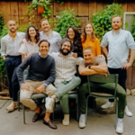 Group of nine people standing and sitting outdoors in front of a wooden wall with plants.