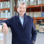 Woman in a blue coat smiling in a warehouse setting, leaning on a wrapped pallet.