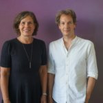 Formal photo of a woman in a black dress and man in a white shirt, standing side by side against a purple wall