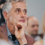 Older man with gray hair smiling gently, resting his chin on his hand in a thoughtful moment.