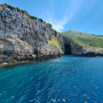 Rocky cliffside with a cave opening next to a blue sea under clear skies.